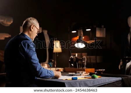 Elderly dressmaker doing needlework in cozy dark atelier shop workspace, checking bespoken clothing drafts. Old couturier looking over sketch drawings, preparing to manufacture garments for customer