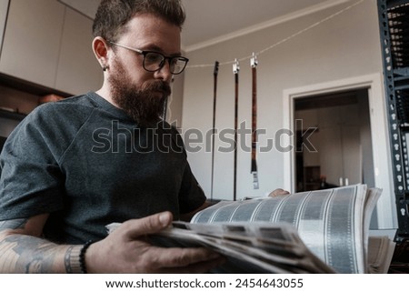 Focused bearded man intensely reviewing film negatives in a modern photography studio with equipment in the background.