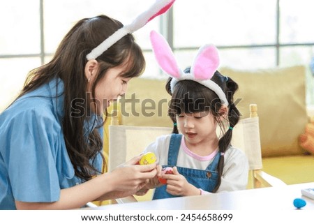 Asian cute little children girl wearing funny bunny ears headbands and young happy mother kiss smile decorating painting eggs while sitting together in living room family preparing for Easter holiday