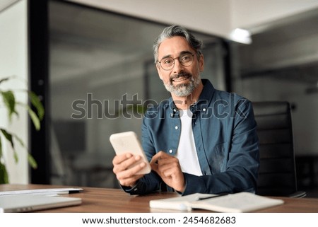 Middle aged businessman looking at camera working holding smartphone. Mature 45 years old business man executive, business owner or entrepreneur sitting at desk using mobile cell phone. Portrait.