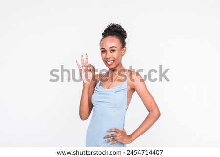Happy multicultural woman in a powder blue dress making an OK gesture, perfectly isolated on a white backdrop.