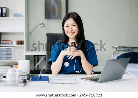 Confident young Asian female doctor in white medical uniform sit at desk working on computer. Smiling use laptop write in medical journal in clinic.
