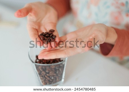 Close-up of girl's hands showing roasted coffee bean with blurred background behind. Shallow depth of field focused on coffee bean. Concept of individual approach to quality control. Royalty-Free Stock Photo #2454780485