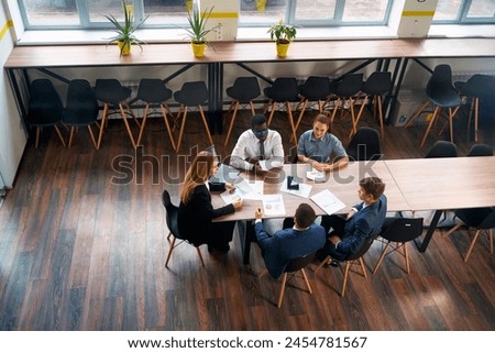 Top view of a diverse group of professionals in a meeting. A female CEO leads a business consultation at a modern office table with multiethnic teammates discussing strategy, growth, and plans.