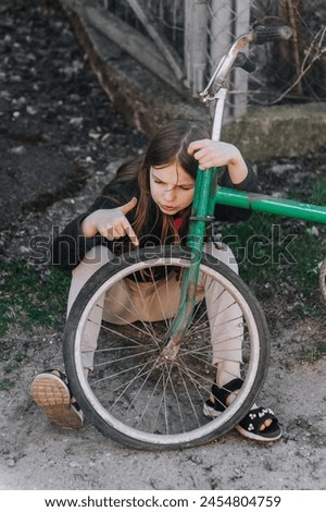 Little beautiful sad teenage girl, dissatisfied upset child showing dislike thumbs down sits near an old bicycle with a broken, punctured wheel tire outdoors. Photography, portrait. Royalty-Free Stock Photo #2454804759