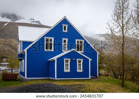 Traditional Icelandic blue residential building with gable roof, white window frames, cladded in corrugated metal sheets in Seydisfjordur town in Iceland with foggy mountains in the background. Royalty-Free Stock Photo #2454831707