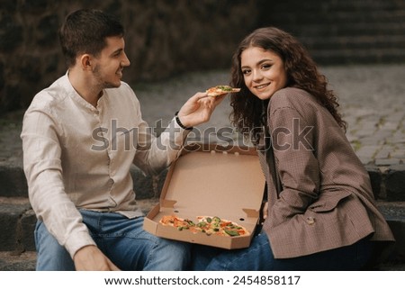 Smiling love couple eating vegan pizza at street on stairs. Handsome man give slice of pizza to woman