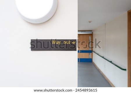 Shallow focus of a Way Out sign seen within an NHS hospital corridor. The door leads to the lobby of a hospital wing.