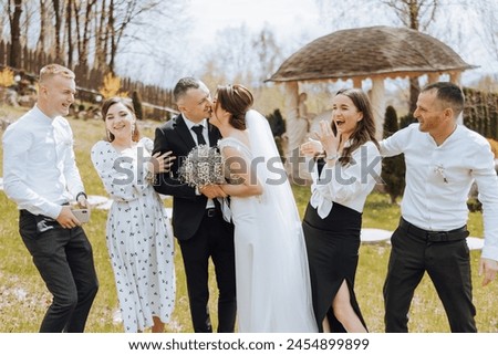 A group of people are posing for a picture, with a bride and groom in the center. The bride is holding a bouquet and the groom is kissing her. The other people in the group are smiling