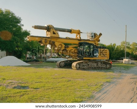 Wide view of a large yellow ROTARY DRILLING RIG in grass and dirt with a pile of small white stones on the left and steel utility poles sections. Paint scrapes and dirt on tracks. Horizontal shot. Royalty-Free Stock Photo #2454908599