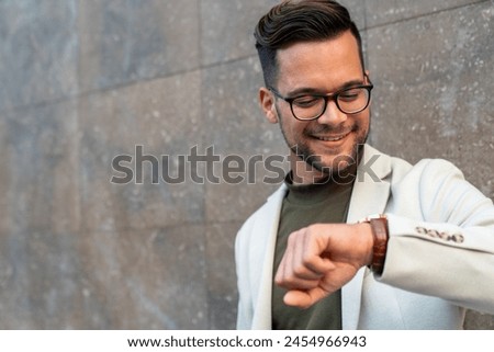 Close up shot of smiling businessman wearing suit and standing against a wall. He is checking time on wrist.