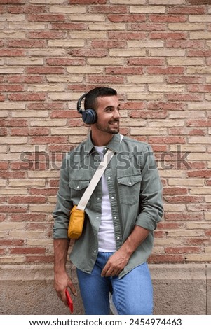Vertical of young adult Caucasian stylish man smiling with wireless headphones holding cell phone leans against brick wall. Attractive male person with confident attitude looking to side outdoors 