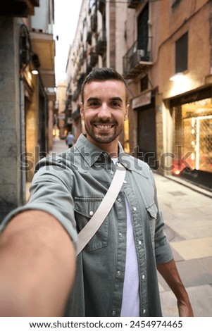 Vertical young happy Caucasian man taking smiling selfie on Madrid touristic city street in background. Millennial handsome tourist takes lighthearted photo outdoors on his vacation trip in Europe 