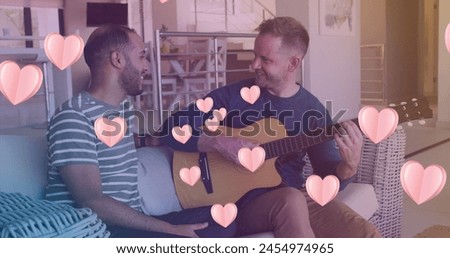 Image of hearts over diverse male couple playing guitar. valentines day and celebration concept digitally generated image.