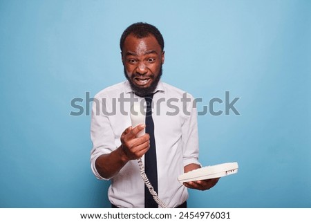 An enraged black businessman yells into the cord phone receiver, arguing with the person on the other end of the line. An anxious African American employee is on the phone with his manager. Royalty-Free Stock Photo #2454976031