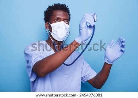 Male healthcare nurse is holding stethoscope to examine cardiology patient heartbeat. African American man wearing gloves and face mask as cardiologist fights coronavirus epidemic. Royalty-Free Stock Photo #2454976081