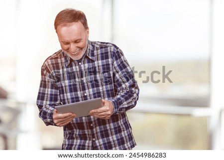 Happy mature aged man is looking down at tablet pc. Bright office background.