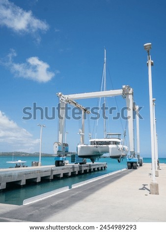Lagoon 42 white Catamaran yacht is suspended in a strong travel lift over a slip and Caribbean blue water on a sunny day with blue sky, white clouds.  Cement foreground and horizon in the background. Royalty-Free Stock Photo #2454989293