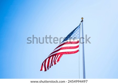 The image shows an American flag flying against a clear blue sky. The flag is waving, indicating a gentle breeze. The flag features the traditional design with alternating red and white stripes and a 