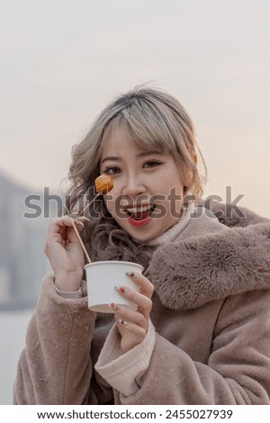 A young woman in winter clothes in her 20s who eats local food fish balls in the evening in a park with a view of buildings and the sea on Kowloon Island, Hong Kong