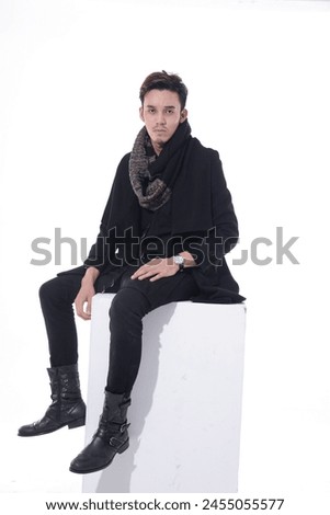 photo of young man wearing coat shirt, jeans sitting cube in studio