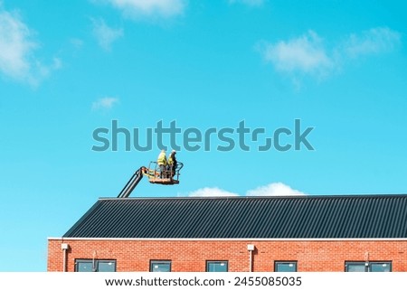 Roofers working off telescopic boom lift while inspecting and replacing roof tiules on a  multistorey residential building against blue sky with space for test. Working at height safely poster concept Royalty-Free Stock Photo #2455085035