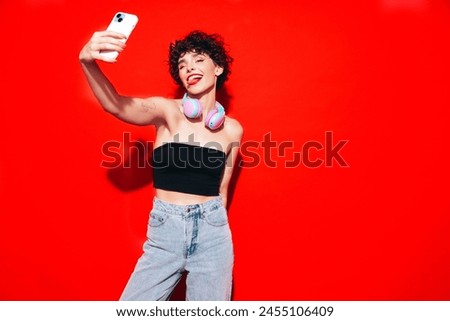 Young beautiful smiling female in trendy summer tank top. Carefree woman posing near red wall in studio with curly hairstyle. Positive model having fun. Listens music, headphones on neck, take selfie