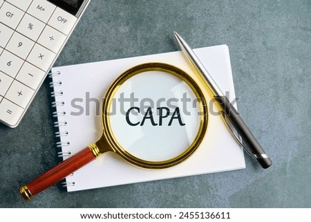 Concept words CAPA, corrective and preventive actions written through a magnifying glass on a notebook in a composition with a calculator, a pen on a gray background