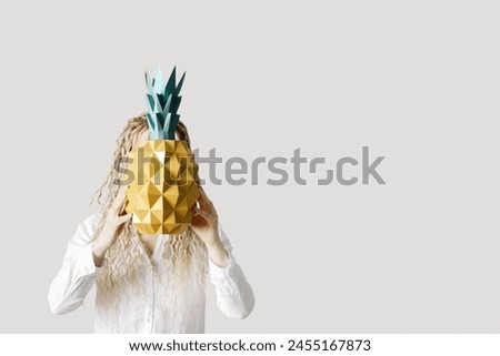 Woman holding paper pineapple in front of face, light grey background, no face trend concept. Young Female with long curly hair dreadlocks hiding behind pineapple. Creative summer vacation concept