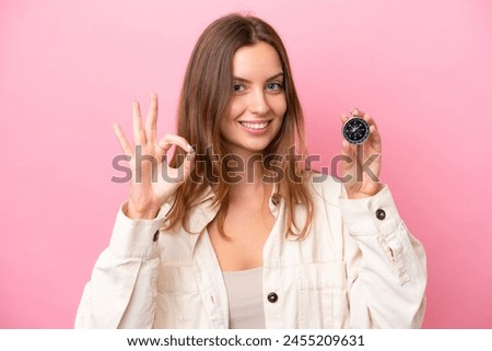 Young caucasian woman holding compass isolated on pink background showing ok sign with fingers