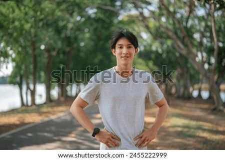 Smiling young asian sportsman man wear sportswear clothes warm up train stand akimbo arms on waist in urban city park, enjoying open air workout in summer Royalty-Free Stock Photo #2455222999