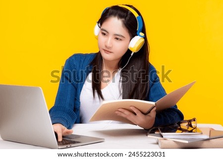 Student young asian woman wearing headphones studying with an open book and laptop on a desk against a yellow background. E-learning Study online with skype teacher, learn language watch webinar Royalty-Free Stock Photo #2455223031