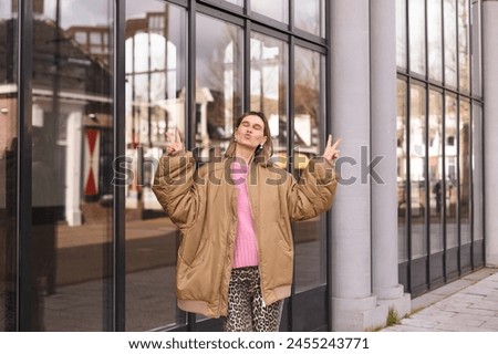 Cool blonde short hair woman in bomber and pink sweater shows peace sign, blows kiss and posing outside. Happy woman walk and posing outdoor near glass building.