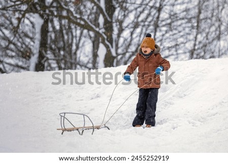 Children's games in the winter outdoors with sledding on a snow mountain. A little boy is calling for help to pull his sled up the hill.