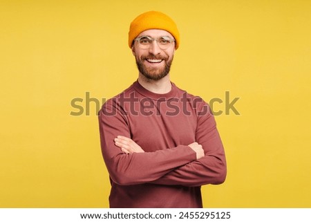 Smiling handsome bearded man, hipster looking at camera and posing arms crossed isolated on yellow background. Happy fashion model posing for pictures, studio shot