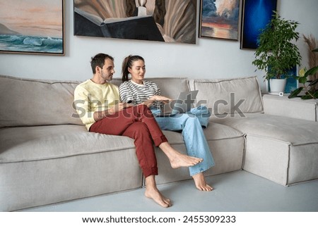 Pleasant family couple artists sits on couch looking at laptop screen browsing internet together. Creative young married spouse web surfing, making purchases online, booking flight tickets for travel. Royalty-Free Stock Photo #2455309233