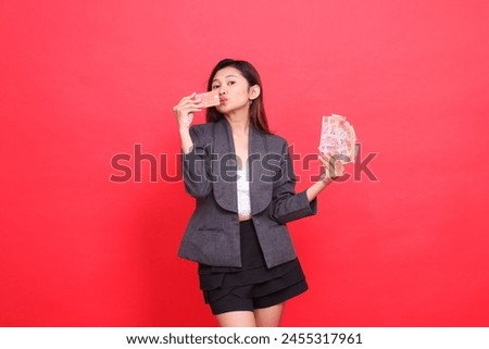gesture of happy indonesia office woman holding while kissing credit cards and money in front of her, wearing a jacket and skirt on a red background. for financial, business and advertising concepts