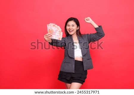 happy indonesia career woman holding a credit card and money in front of a sign clenching her fist to the side wearing a jacket and skirt on a red background. for transaction, business and advertising