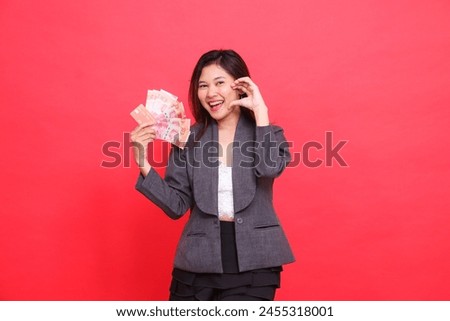 The expression of a happy indonesia office woman holding a credit, debit card and money while showing love to the camera wearing a gray jacket and skirt on a red background. for business, advertising