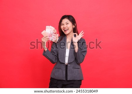 The expression of a happy Asian office woman holding a credit, debit card and money while signing okay (good) to the camera wearing a gray jacket and red skirt. for transaction, business, advertising