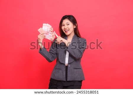 Asian office woman smiling expression holding credit, debit card and money while pointing top right at the camera wearing gray jacket and red skirt. for transaction, business and advertising concepts