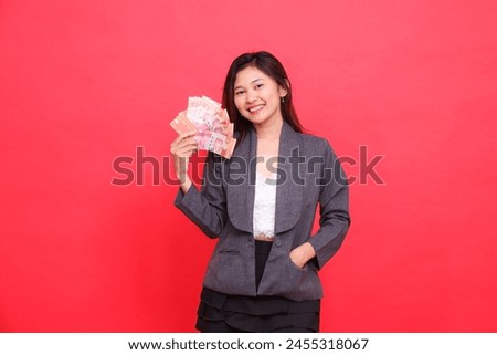 Expression of cheerful indonesia office woman holding debit credit card and money upright and in pocket looking at camera wearing gray jacket and red skirt. for transaction, business and advertising