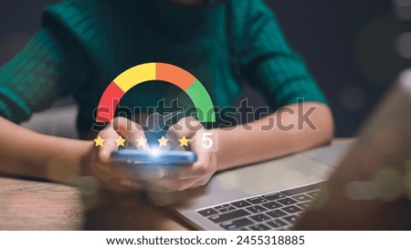 A woman is holding a cell phone with a green screen that says 5. She is sitting at a table with a laptop in front of her