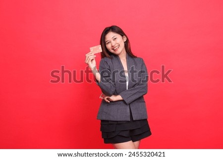 Expression of cheerful indonesia businesswoman with arms crossed and holding debit credit card wearing jacket and skirt on red background. for financial, business and advertising concepts