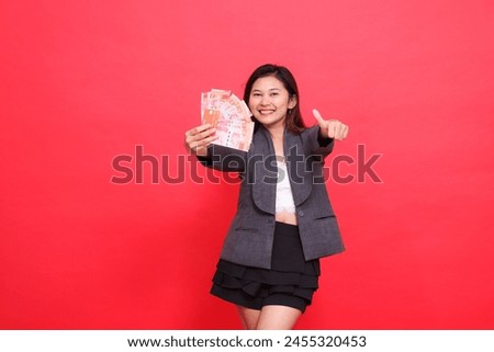 gesture of a happy indonesia career woman holding a credit card and money in front of a thumbs up sign wearing a jacket and skirt on a red background. for transaction, business and advertising
