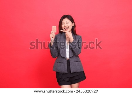 Cheerful candid expression of Asian office girl in hand holding debit credit card while pointing at it wearing jacket and skirt on red background. for financial, business and advertising concepts
