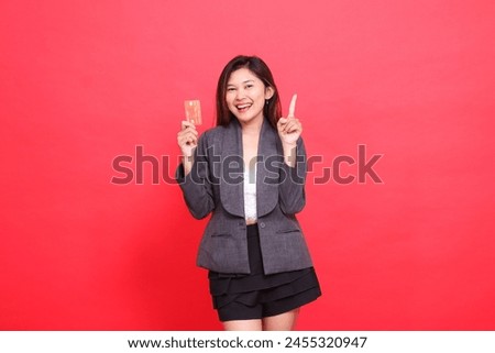 Cheerful indonesia office woman's expression into the camera holding a debit credit card while pointing upwards wearing a jacket and skirt on a red background. for financial, business and advertising