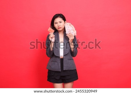 gesture of surprised indonesia office woman holding credit card and money in head area wearing jacket and skirt on red background. for transaction, business and advertising concepts