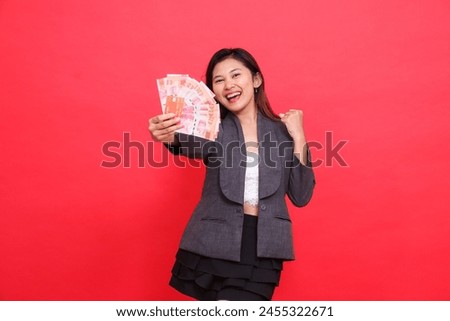 the expression of a cheerful indonesia director woman holding a credit card and money in front of her clenched fist at the camera wearing a jacket and skirt on a red background. for business advert