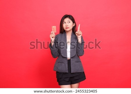 chinese office woman's expression shocked with her hand holding a debit credit card while pointing upwards, wearing a jacket and skirt on a red background. for financial, business and advertising